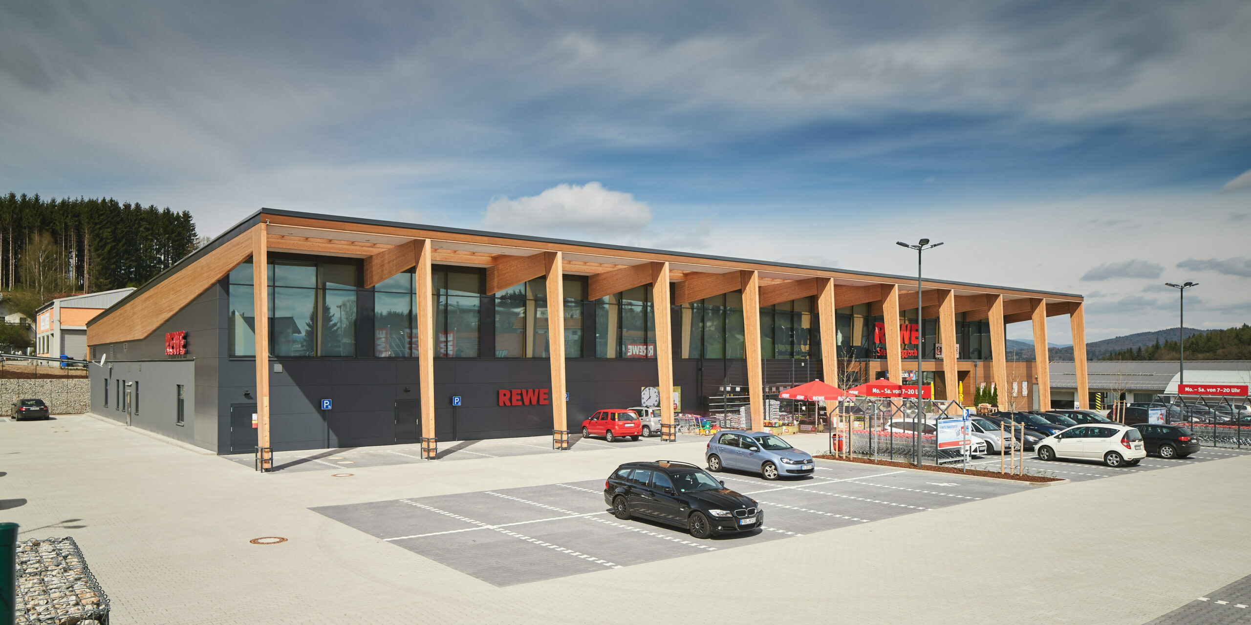 Beispiel eines Green Building REWE-Markts bei Tageslicht. Das Gebäude ist grau und hat einen überdachten Vorbau aus Holz. Vor dem Gebäude ist ein Parkplatz, auf dem einige wenige Autos stehen.