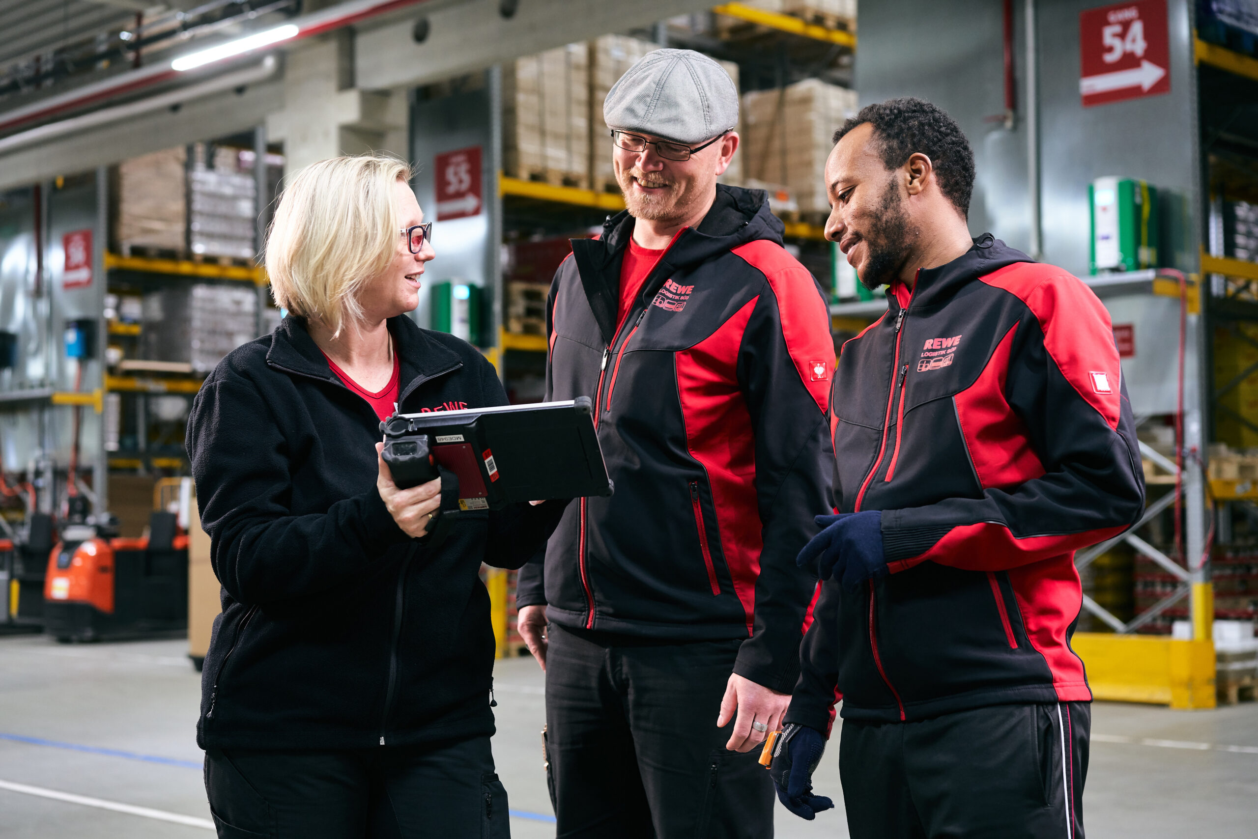 Drei Logistikmitarbeiter, die im Zentrallager stehen, eine Frau und zwei Männer. Die Frau hält ein Tablet in der Hand und zeigt ihren Kollegen den Bildschirm.