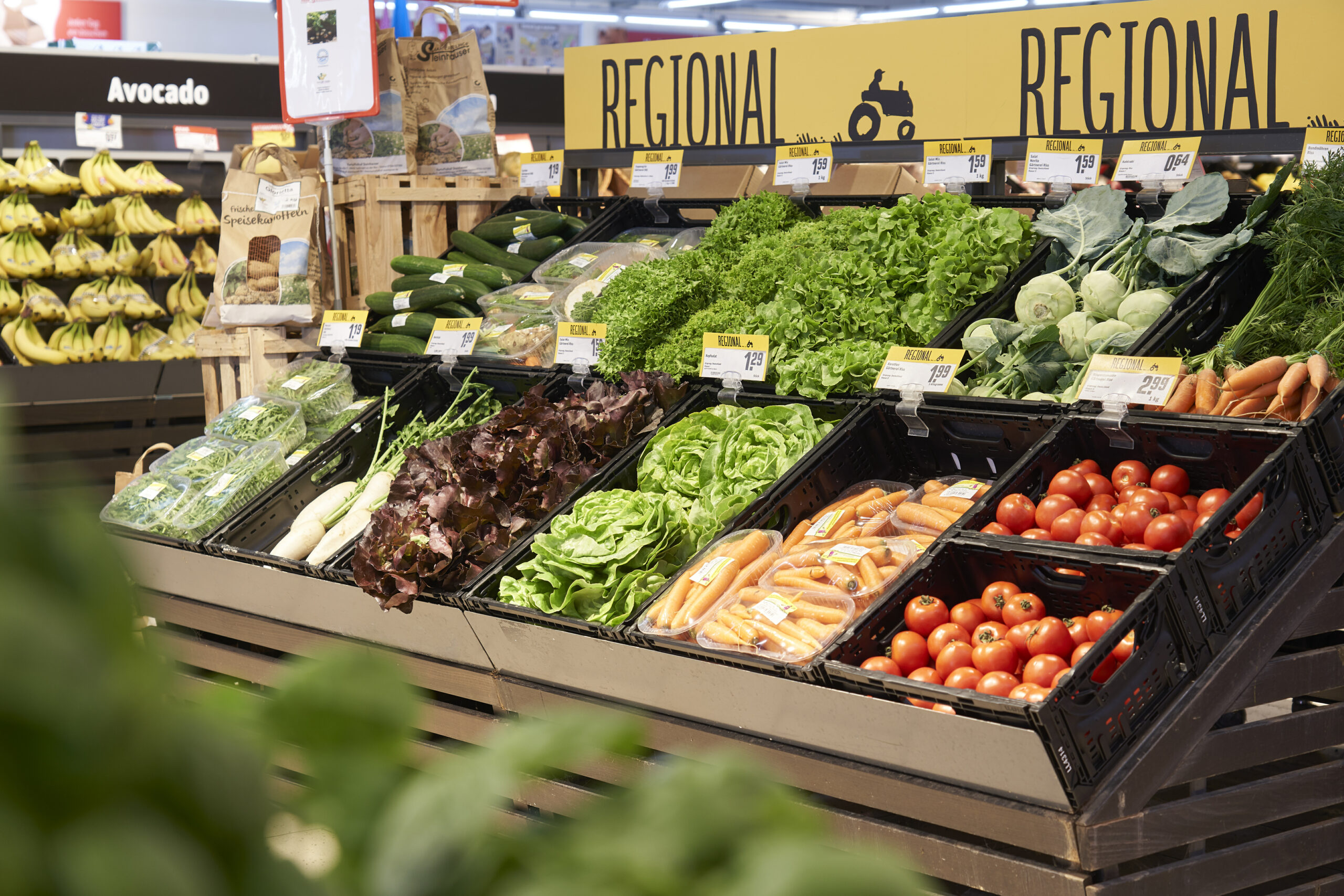 Wareninsel mit einem Schild, dass auf regionale Ware hinweist. Auf der Wareninsel sind Kisten mit Gemüse, darunter zum Beispiel Kohlrabi, Tomaten, Karotten, Salatköpfe und Gurken.