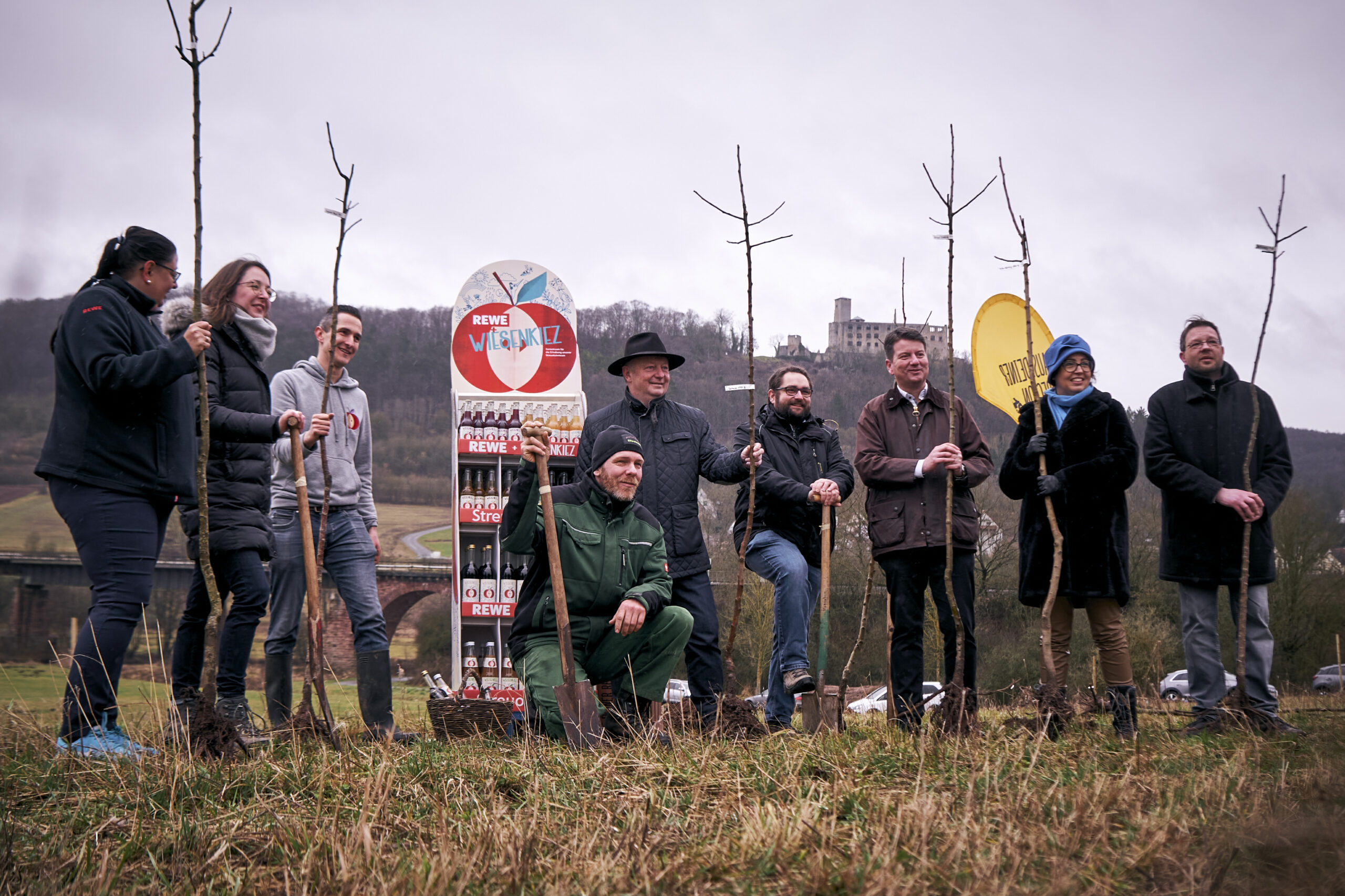 Neun Personen auf einem Feld, die Bäume pflanzen. Im Hintergrund steht ein Aufsteller mit Getränken.