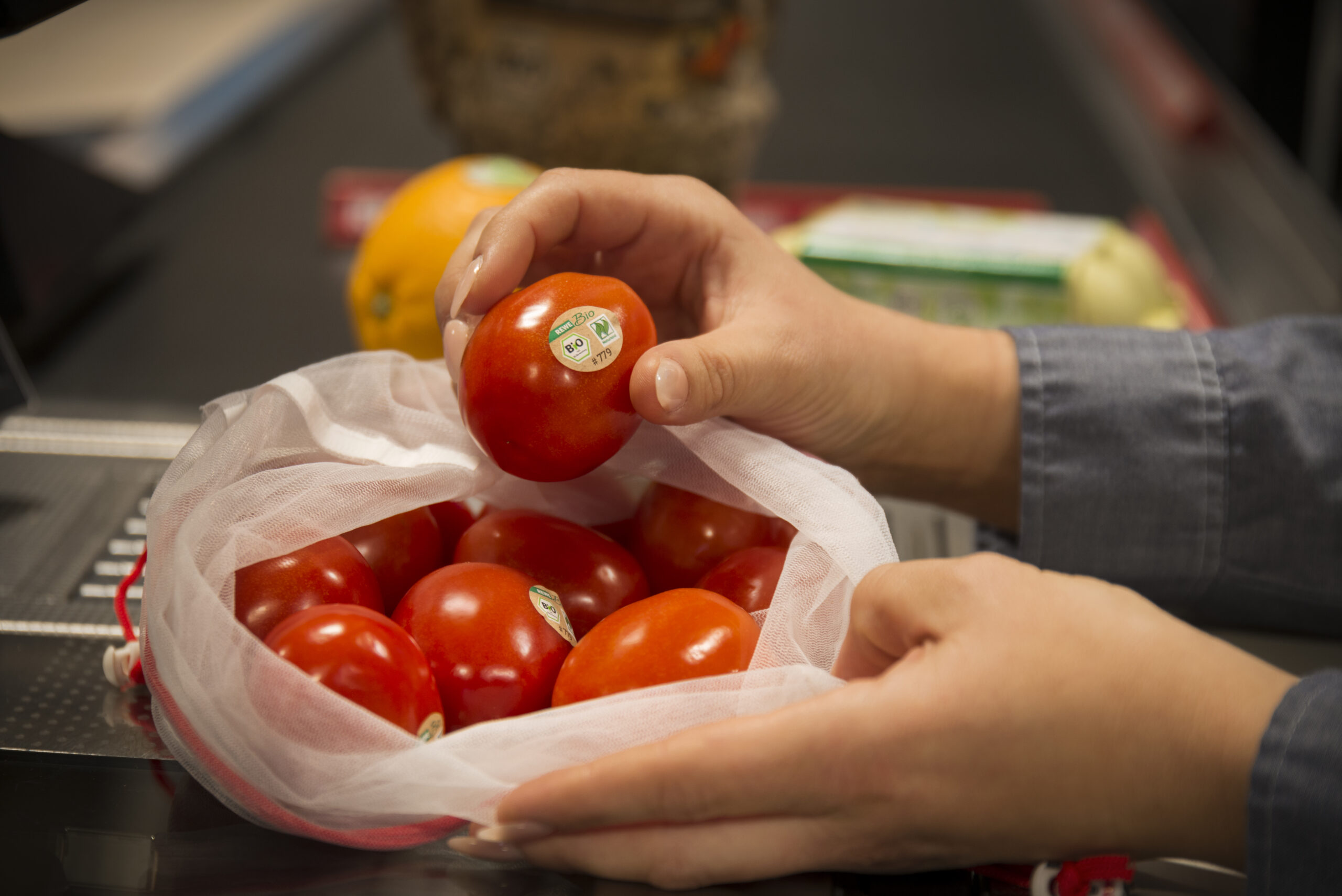 Stoffsäckchen mit Tomaten auf der Kassenfläche. Die Verkäuferin hält eine der Tomaten in der Hand. Die Tomaten sind mit einem Bio-Aufkleber versehen.